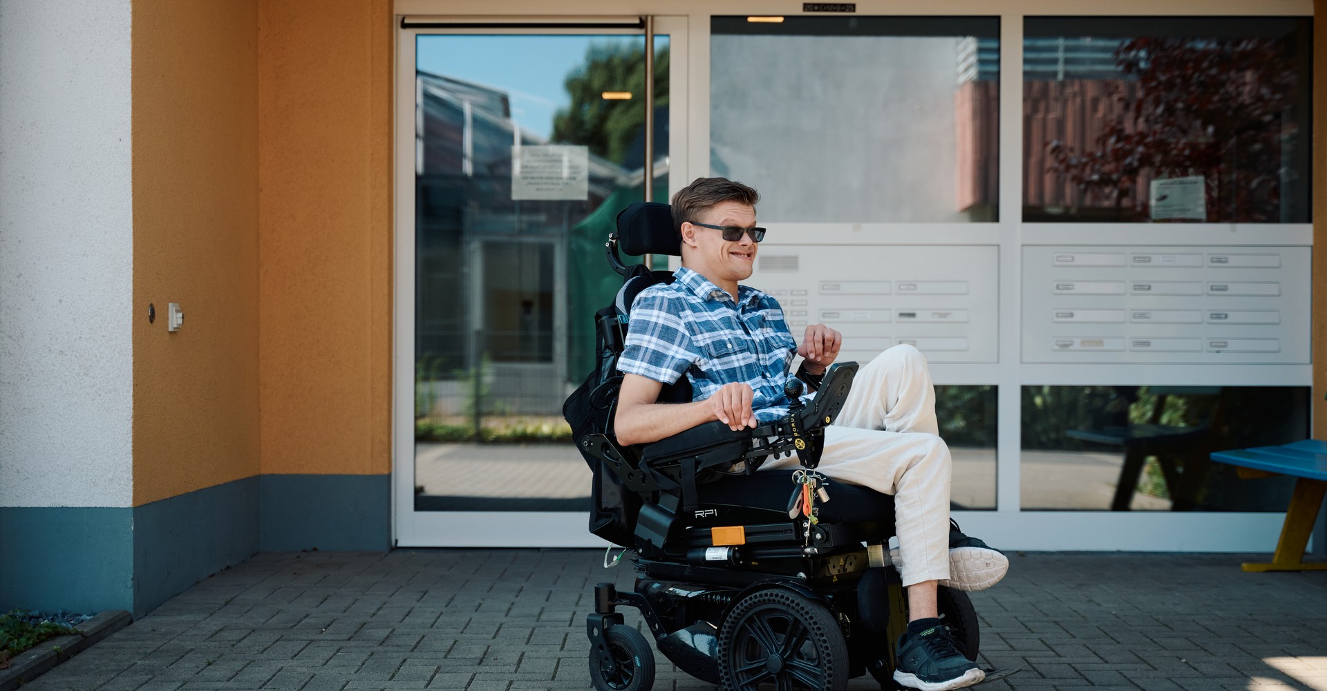 Ein junger Mann im sitzt im elektrischen Rollstuhl vor dem Eingangsbereich eines Mehrfamilienhauses.