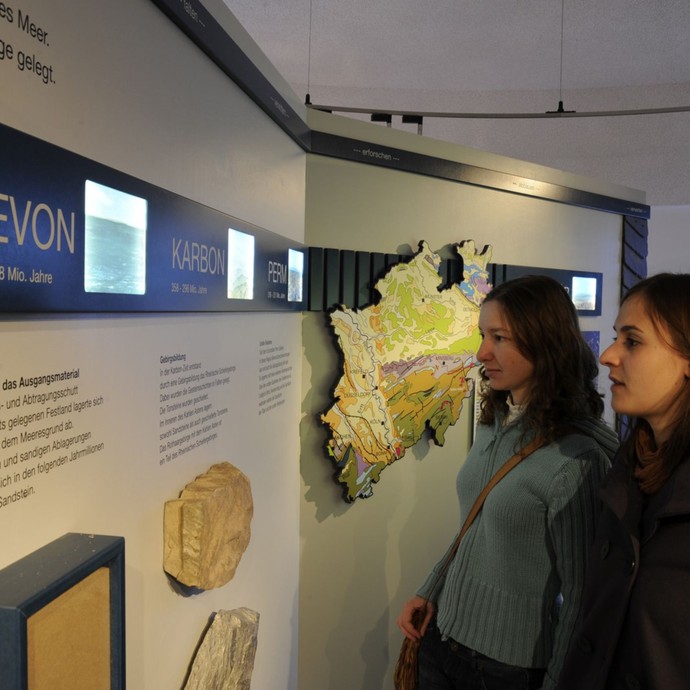 Zwei Mädchen schauen auf eine Ausstellungswand mit Text, Bildschirmen und Gestein (vergrößerte Bildansicht wird geöffnet)