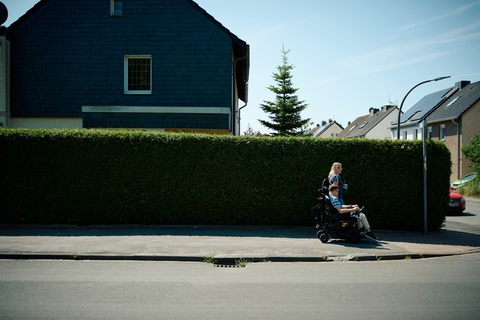 Eine junge Frau geht neben einem jungen Mann im Rollstuhl. Im Hintergrund eine Hecke, dahinter ein Haus.