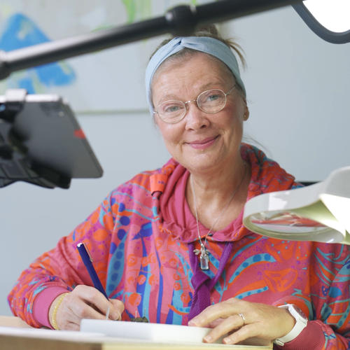 Ein Frau sitzt am Zeichentisch und lächelt in die Kamera.