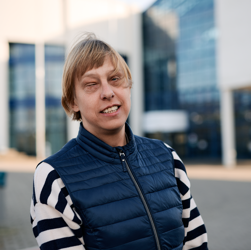 Sabrina Führer mit blonden Haaren, blauer Weste und blaue-weiß gestreiftem Pullover lächelt in die Kamera, Glasfassade im Hintergrund