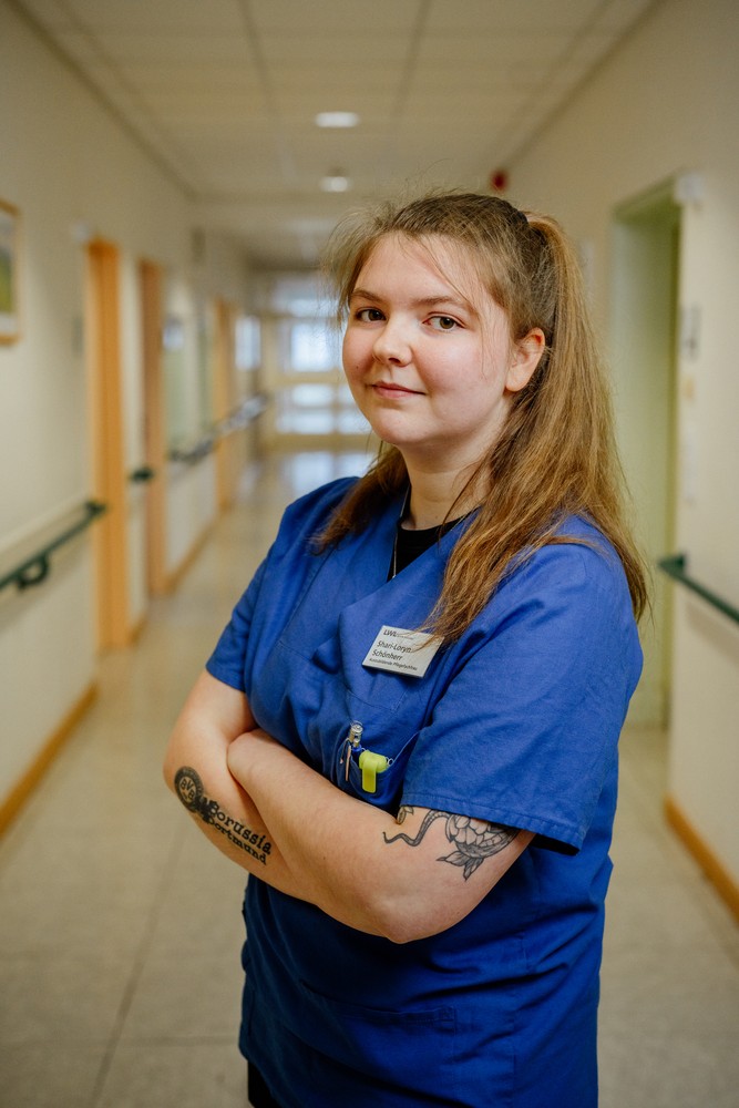 Porträt einer jungen Frau im blauen Pfleger:innen-Oberteil. Sie steht in einem Krankenhausflur.