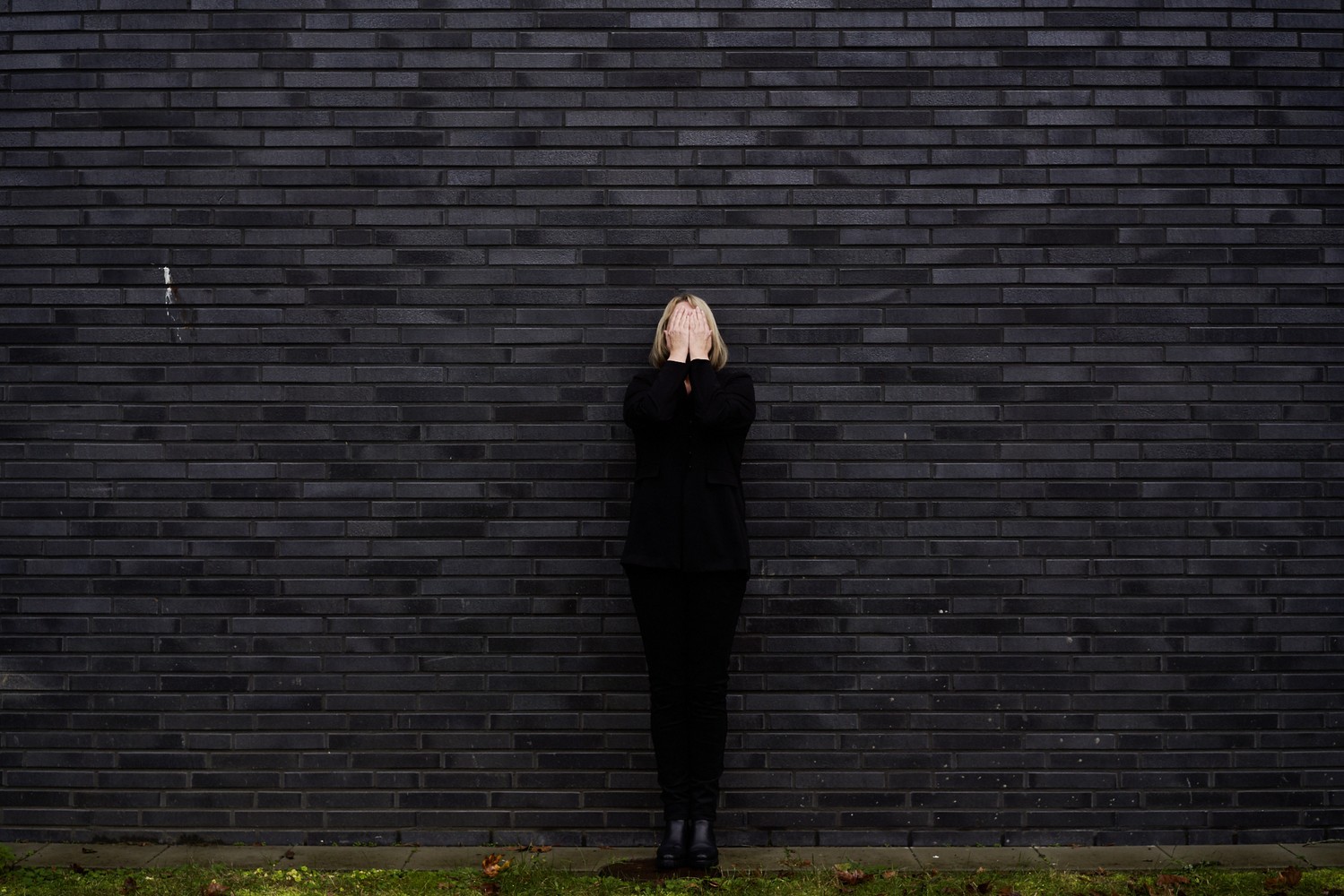 Eine Frau in schwarzer Kleidung steht vor einer schwarzen Wand und hält die Hände vor dem Gesicht.
