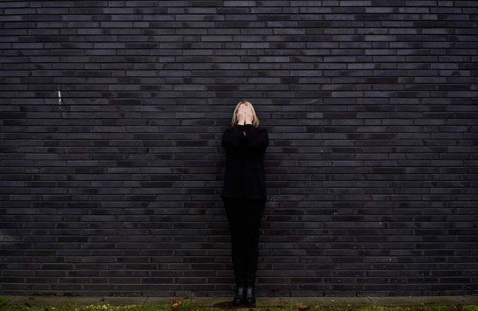 Eine schwarz gekleidete Frau steht vor einer schwarzen Mauer und hält sich beide Hände flach vor ihr Gesicht.