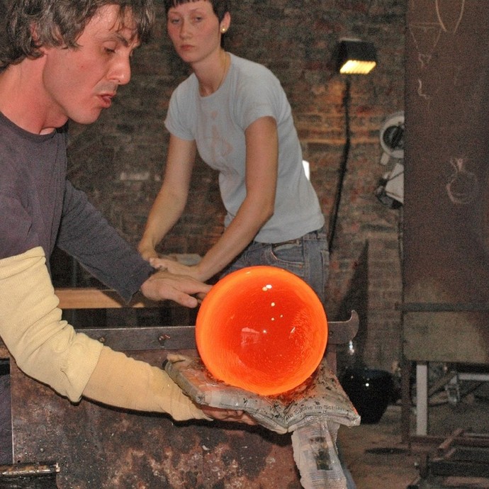 Ein Mann fertigt eine große orangene Glaskugel an. Im Hintergrund steht eine Frau. (vergrößerte Bildansicht wird geöffnet)