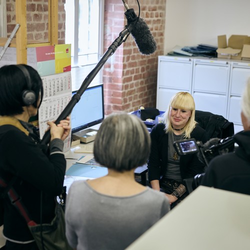 Filmteam mit Kamera und Tonangel filmt eine junge Frau mit blondem Haar an ihrem Schreibtisch.