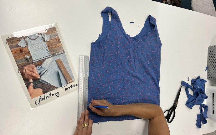 Person zeichnet mit Lineal und Stift auf einem blauen ärmellosen Oberteil, daneben liegen Schere, Stoffreste und eine Anleitung mit dem Schriftzug 'Anleitung basteln'.