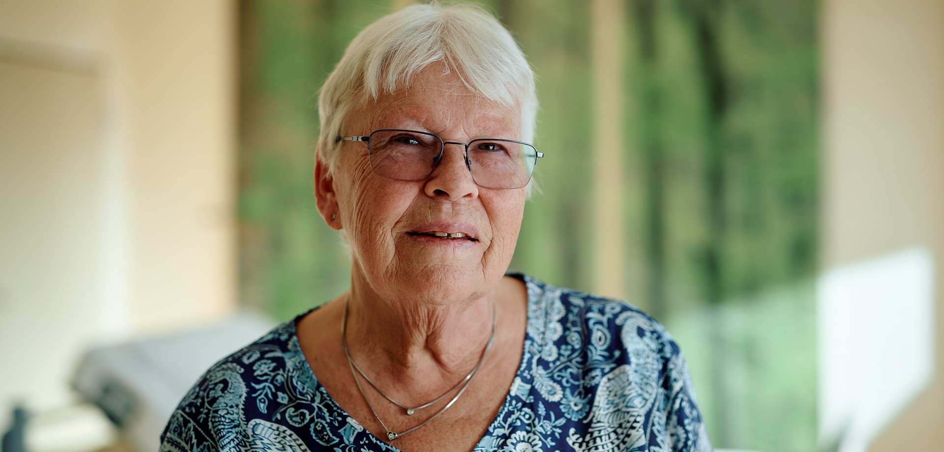 Im Vordergrund eine Frau, dahinter ein Pflegebett und grüne Gardinen