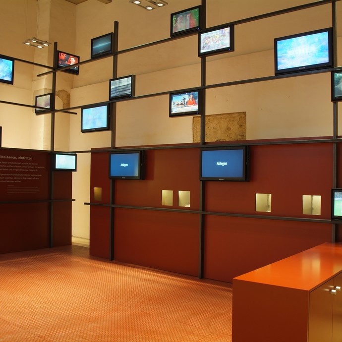 Ein Ausstellungsraum zeigt viele kleine Bildschirme, die an einer Art Holzgitter befestigt sind. (öffnet vergrößerte Bildansicht)