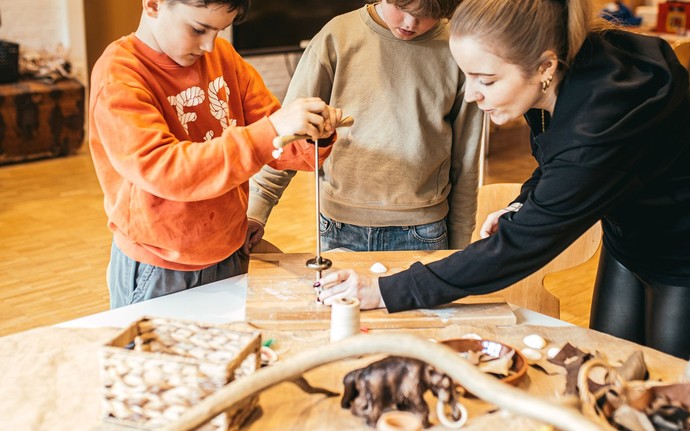 Zwei Schulkinder und eine Frau stehen an einem Tisch und bohren ein Loch in eine Muschel.