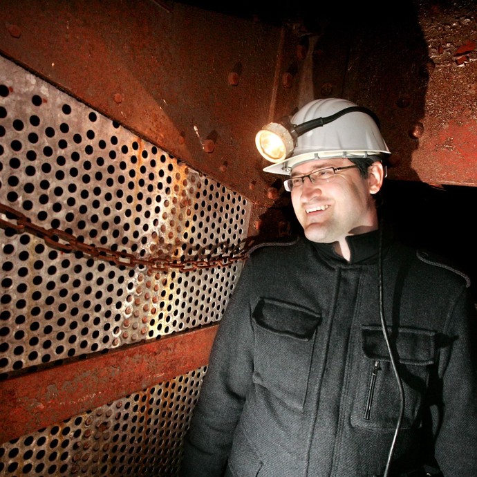 Ein Mann steht mit Schutzhelm und Stirnlampe in einem dunklen Schacht und lächelt (vergrößerte Bildansicht wird geöffnet)